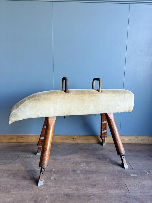 Vintage pommel horse grey padded upholstery with red accents and wooden legs, aged patina.